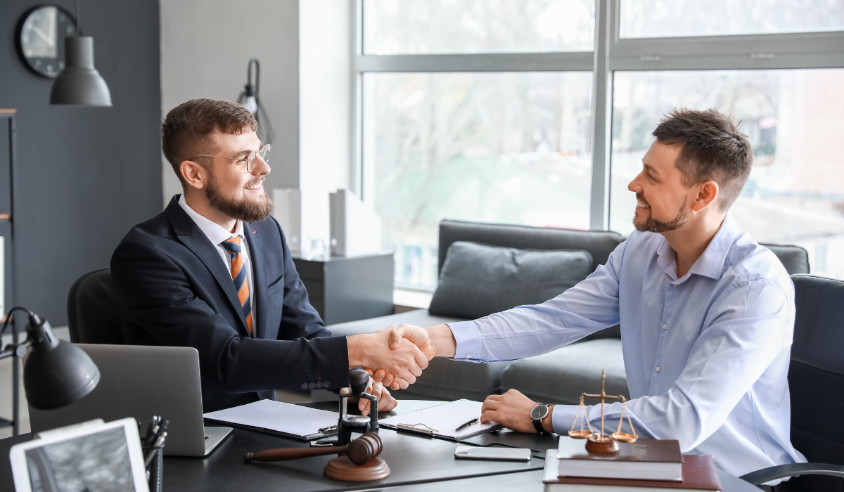 A lawyer meets with an interpreter offering legal language services in a bright office. They shake hands, smiling with legal scales, electronics, and paperwork on the desk.