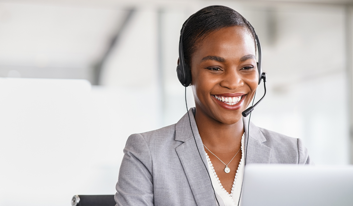 A woman wearing a headset and a gray blazer smiles while working at a laptop in a bright office setting as she connects for a video remote interpreting session.