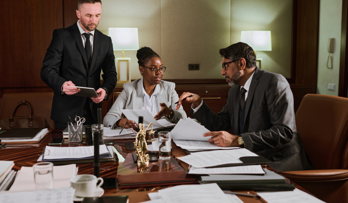 Three professionals in formal attire at a conference table covered with papers, office supplies, and a justice scale review legal documents and discuss contracts with an interpreter providing legal translation services.