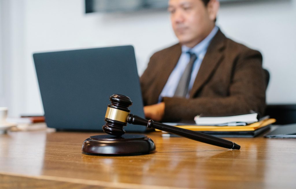 A wooden gavel rests on a desk in front of a man in a suit working on a laptop, with documents, a pen, and a stenographer’s notes nearby.
