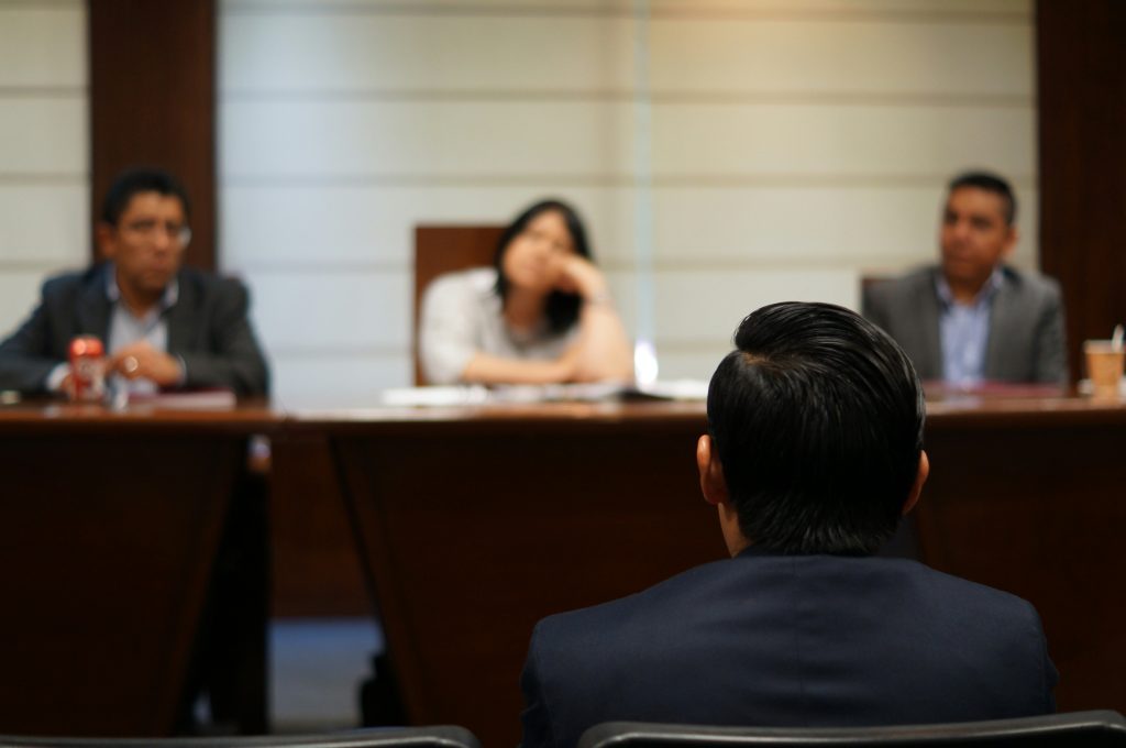 A person sits facing three people at a table in a formal setting, as if participating in an interview or jury selection process.